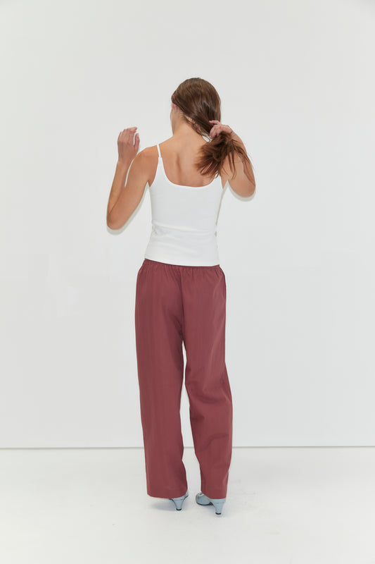A woman stands facing a white wall, holding her brown hair in a ponytail. She wears the Rib Jersey Tank in White from Deiji Studios, loose maroon pants, and light blue heels.