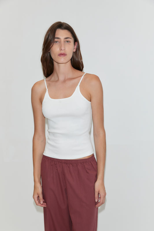 A woman with long brown hair wears the Rib Jersey Tank in White by Deiji Studios and maroon pants as she stands against a plain light-colored background.