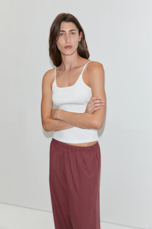 A person with long brown hair wearing a white tank top and the Ease Trouser - Rust Stripe stands against a plain white background with arms crossed, looking at the camera.