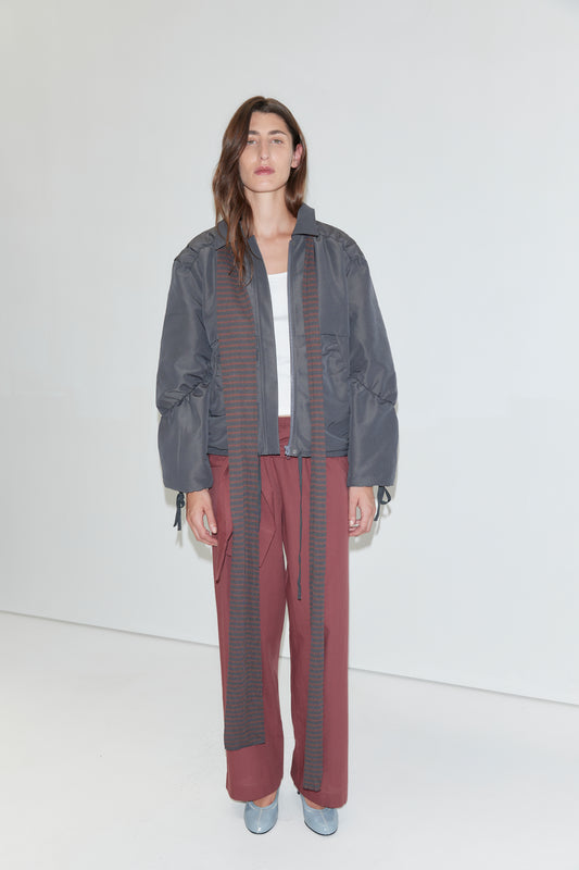 A person stands against a white background wearing a gray bomber jacket, a white top, the Ease Trouser - Rust Stripe from Deiji Studios, a long striped scarf, and light blue shoes. They have long brown hair and a neutral expression.