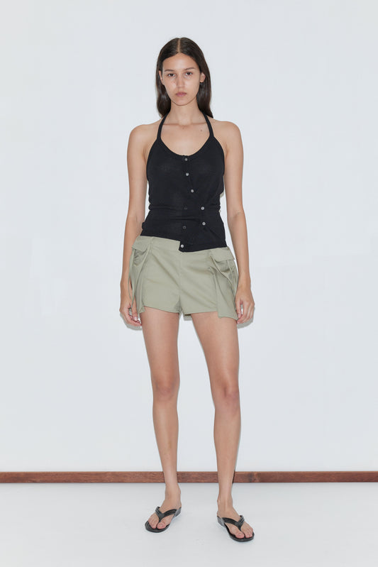 A woman with straight dark hair and a neutral expression stands by a plain white wall, wearing the Cross Top - Black, light olive green shorts with side ties, and black flip-flops.
