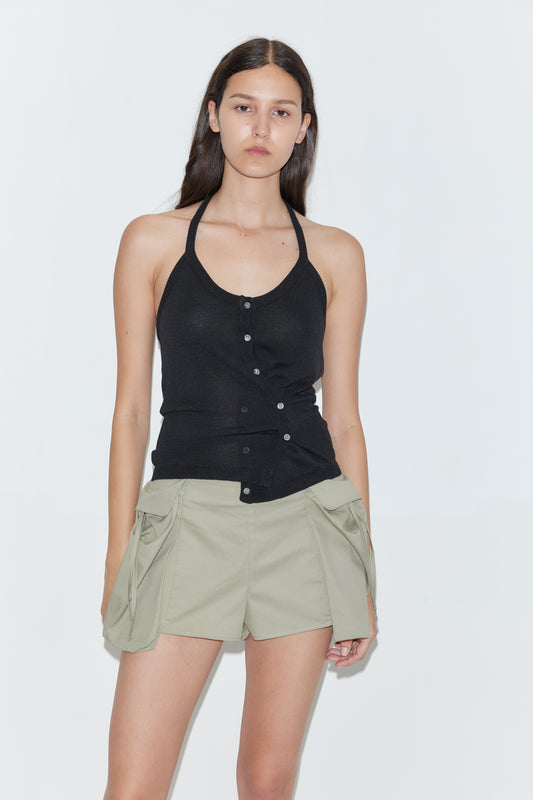 A woman with long dark hair wears the Cross Top - Black, a sleeveless knit halter top with buttons, paired with light khaki shorts featuring large side pockets, standing against a plain white background.
