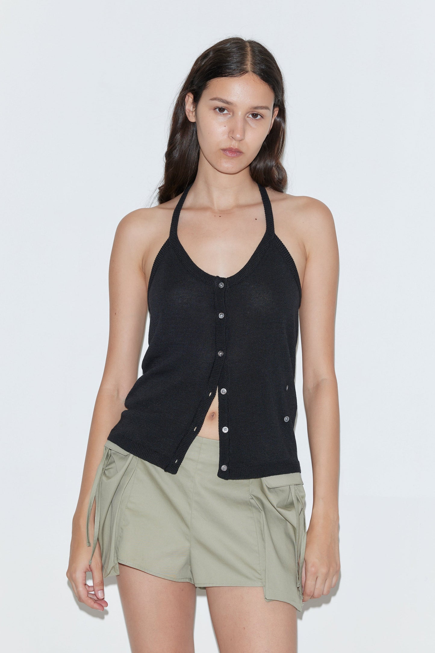 A woman with long, dark hair and a neutral expression wears the Cross Top - Black by Deiji Studios and light green shorts with pocket flaps and side ties, creating an effortlessly chic look against a plain white background.