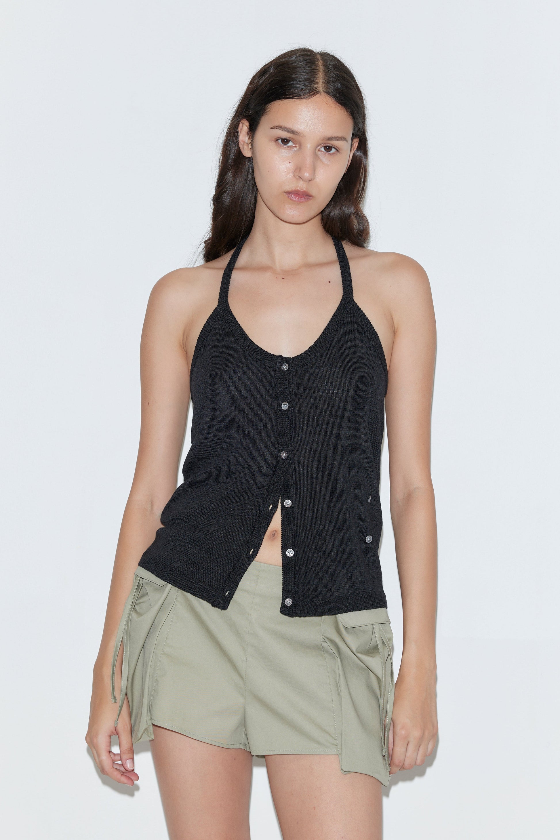 A woman with long, dark hair and a neutral expression wears the Cross Top - Black by Deiji Studios and light green shorts with pocket flaps and side ties, creating an effortlessly chic look against a plain white background.