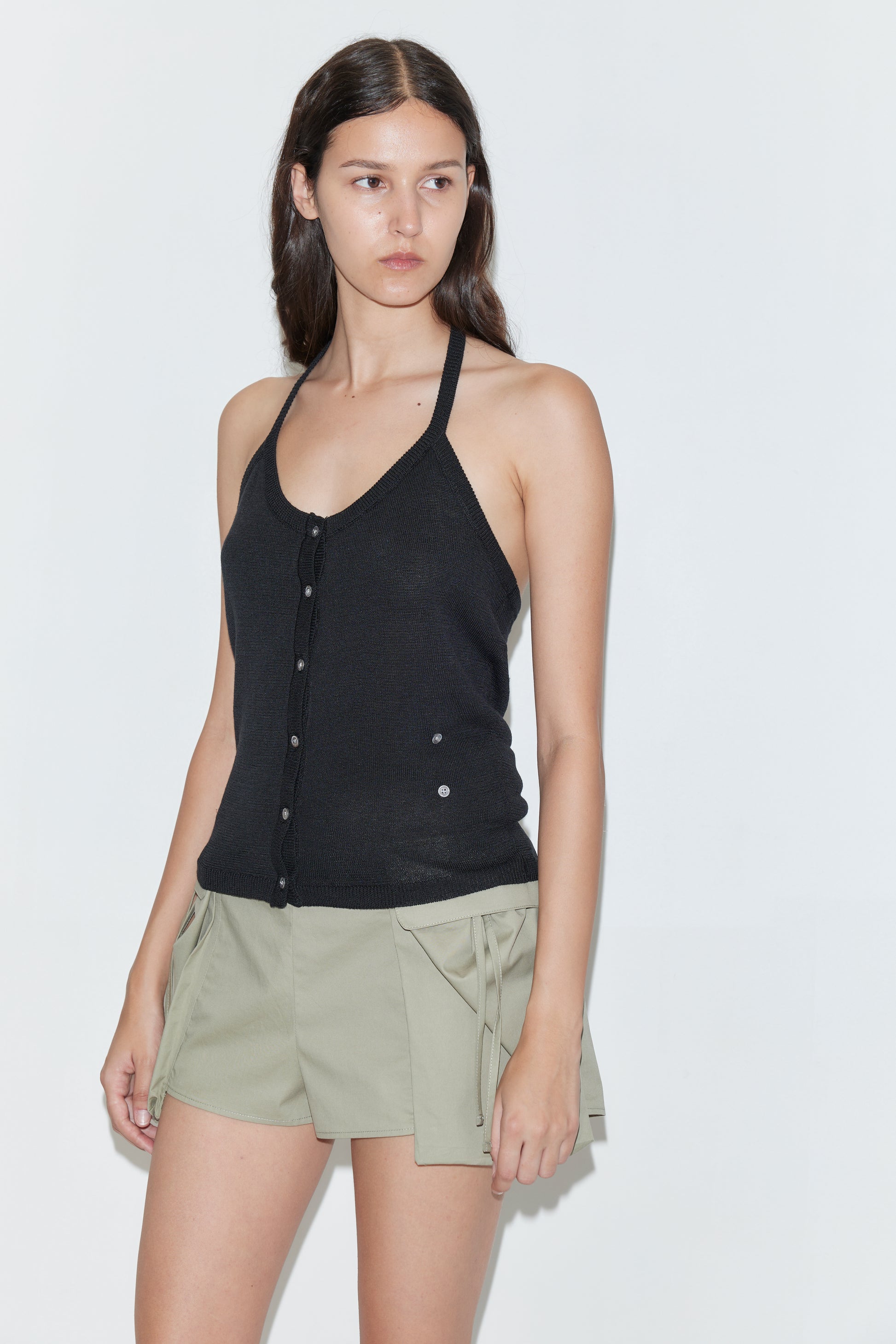 A woman with long dark hair wears the Cross Top - Black by Deiji Studios and light khaki shorts with large front pockets, standing against a plain white background.