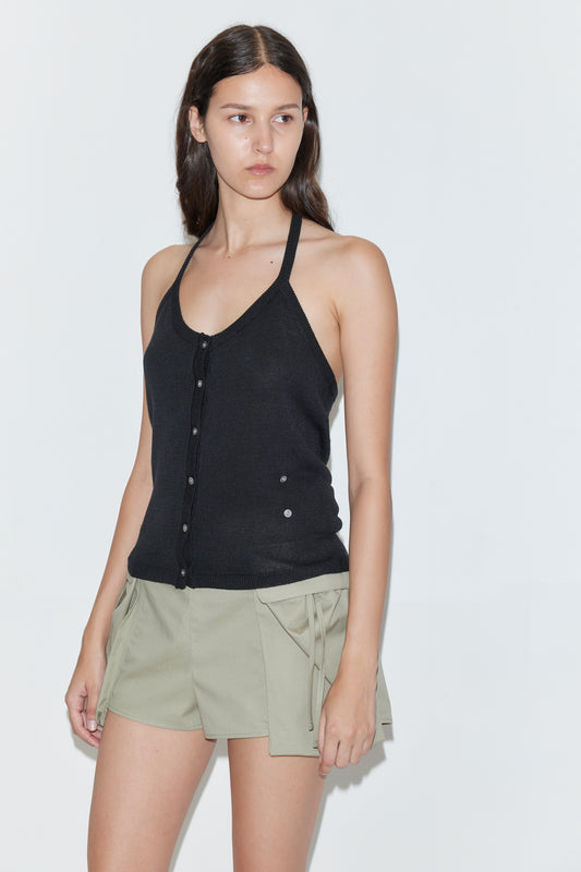 A woman with long dark hair wears the Cross Top - Black by Deiji Studios and light khaki shorts with large front pockets, standing against a plain white background.
