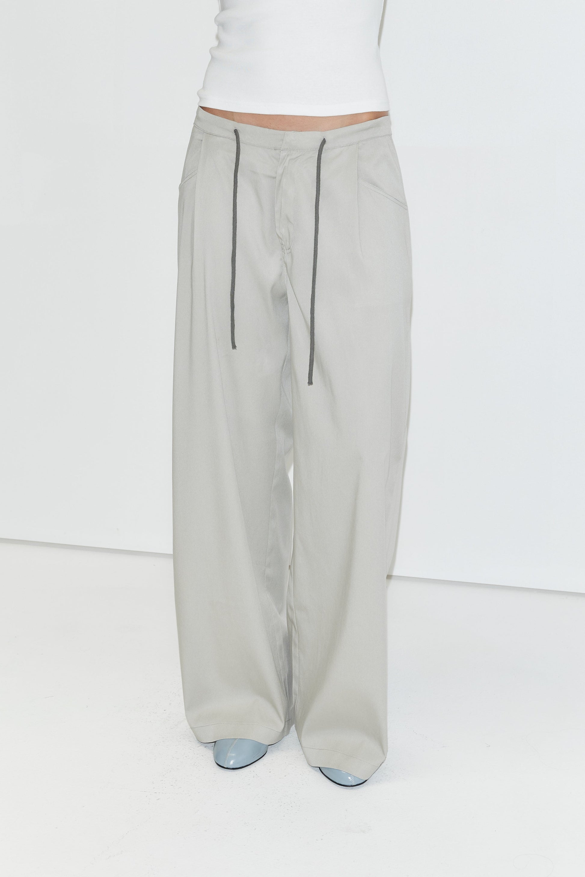 A person wears a white tank top tucked into the Tencel Pant - Grey by Deiji Studios, which features a comfortable drawstring waist and a loose, wide-leg fit. The look is completed with light blue pointed-toe shoes against a plain white studio background.