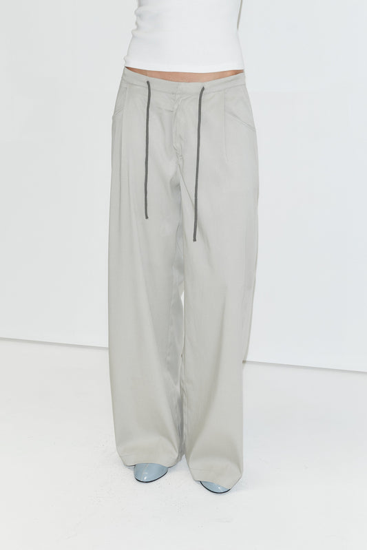 A person wears a white tank top tucked into the Tencel Pant - Grey by Deiji Studios, which features a comfortable drawstring waist and a loose, wide-leg fit. The look is completed with light blue pointed-toe shoes against a plain white studio background.