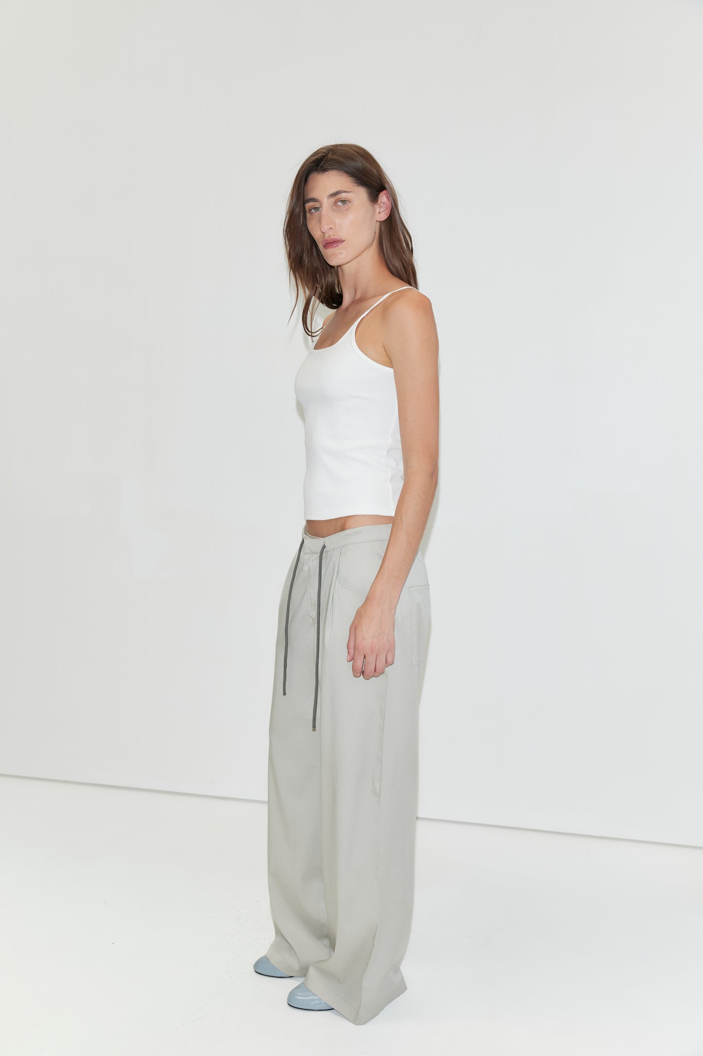 A woman with long brown hair wears a white tank top, the Tencel Pant - Grey from Deiji Studios—loose light gray wide-leg pants with drawstrings—and blue shoes, standing against a plain white background.