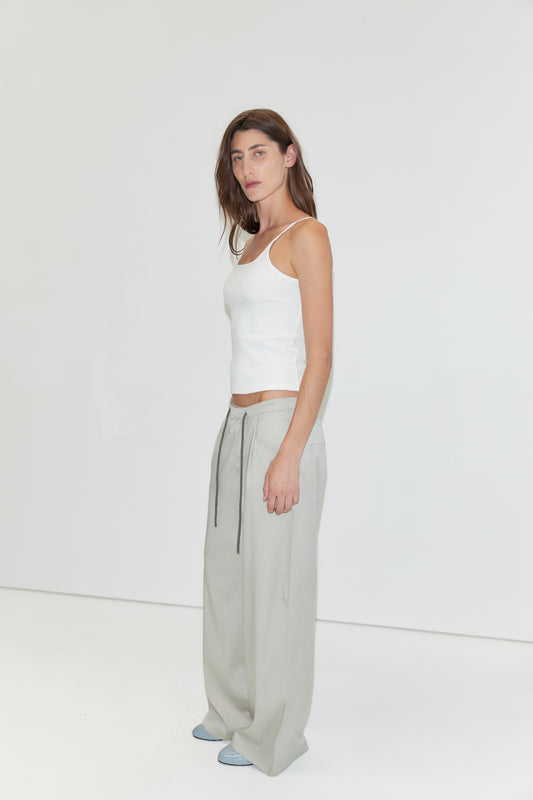 A woman with long brown hair wears a white tank top, the Tencel Pant - Grey from Deiji Studios—loose light gray wide-leg pants with drawstrings—and blue shoes, standing against a plain white background.