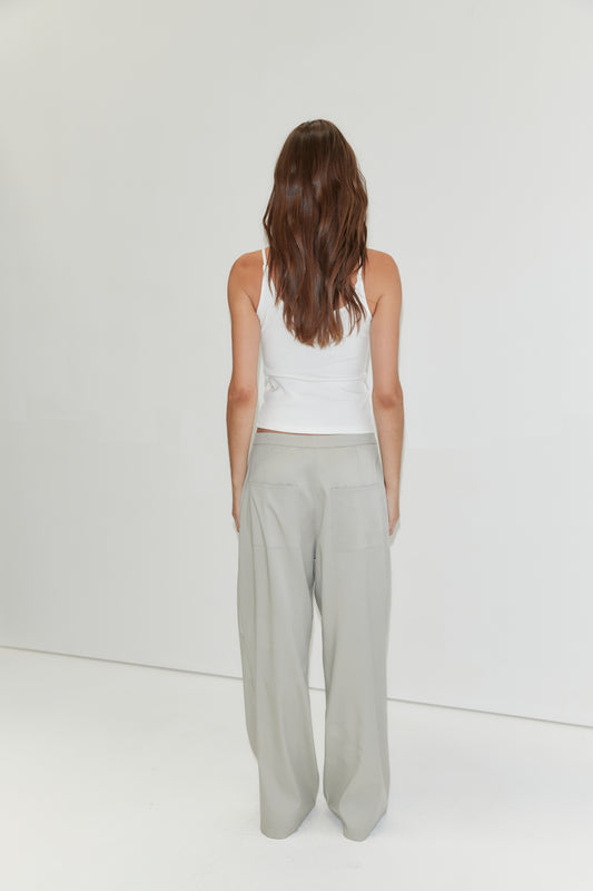 A person with long brown hair stands facing away, wearing a white tank top and the Tencel Pant - Grey from Deiji Studios, against a plain white background.