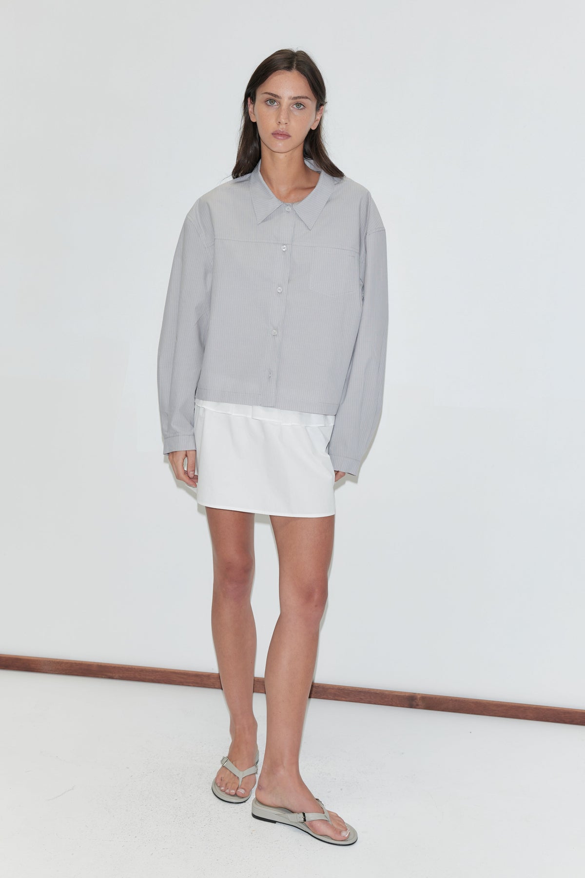 A woman with long dark hair stands against a plain white background wearing the Flounce Layer Mini Skirt - White by Deiji Studios, a white low-rise mini skirt with a flounce waist, with a pale grey striped boxy shirt and flip-flops.