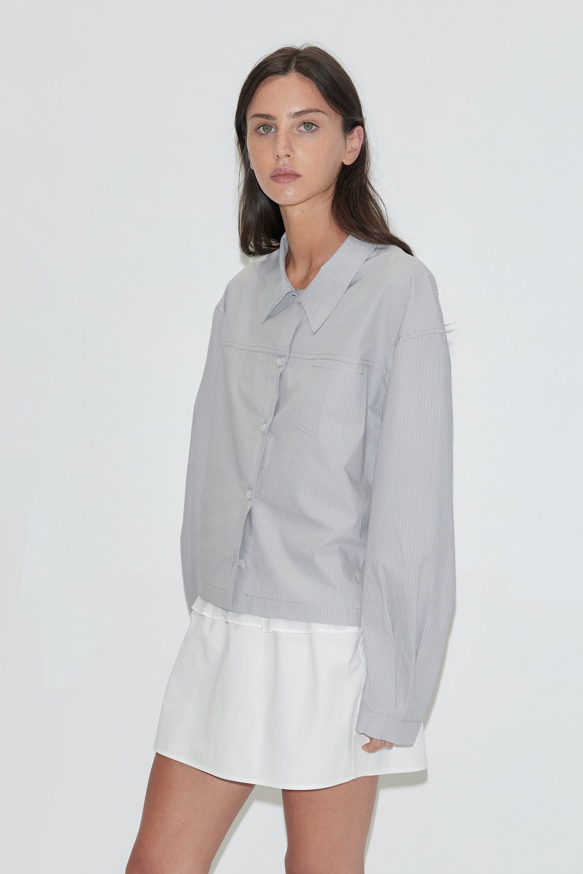 A woman with long dark hair stands against a plain white background wearing the Boxy Shirt - Cloud Stripe by Deiji Studios, a pale grey-blue fine-striped boxy shirt, over a white flounce mini skirt with grey flip-flops.