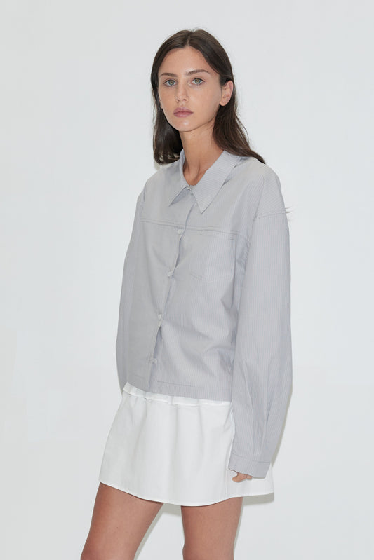 A woman with long dark hair stands against a plain white background wearing the Boxy Shirt - Cloud Stripe by Deiji Studios, a pale grey-blue fine-striped boxy shirt, over a white flounce mini skirt with grey flip-flops.