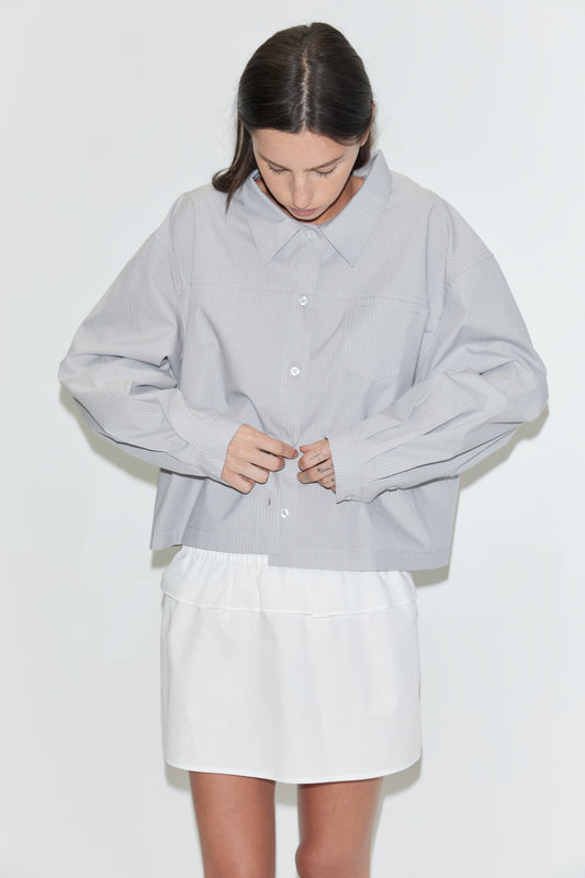 A cropped view of a woman looking down, wearing the Flounce Layer Mini Skirt - White by Deiji Studios, a white low-rise mini skirt with a flounce layer at the waist, and a pale grey striped boxy shirt against a plain white background.