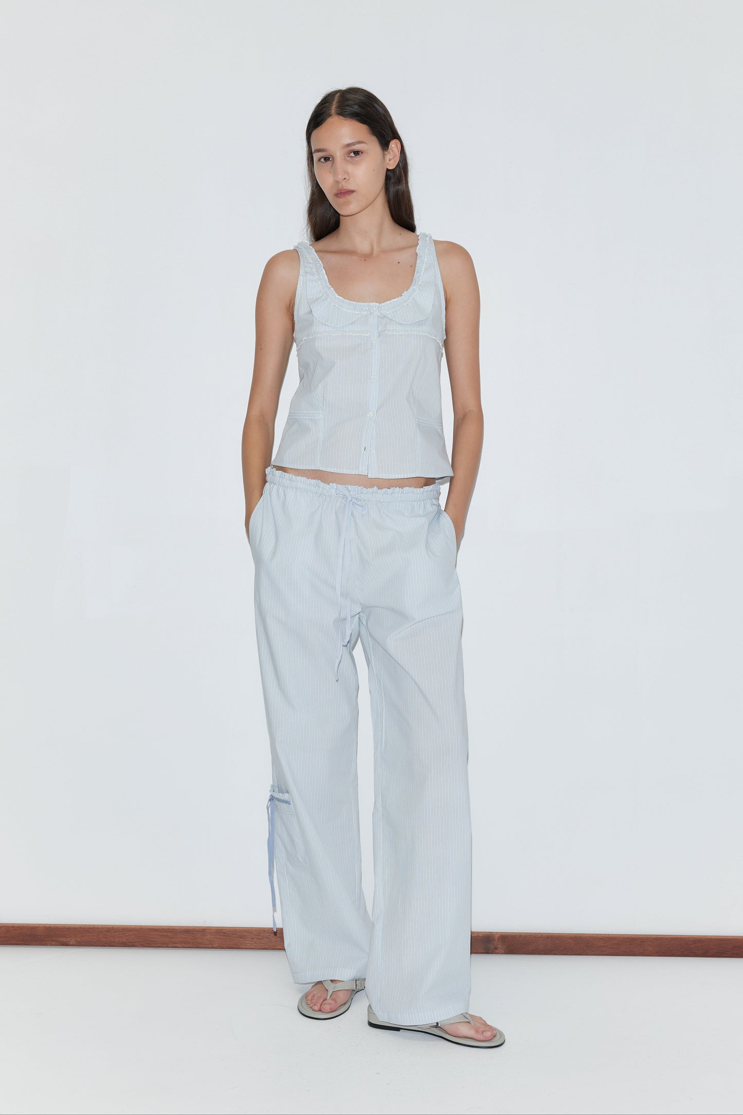 A woman stands against a plain white wall wearing the Pintuck Collar Set - Mist Stripe, featuring a light blue top and matching wide-leg pants. She has her hands in her pockets, pairs the set with sandals, and maintains a neutral expression.