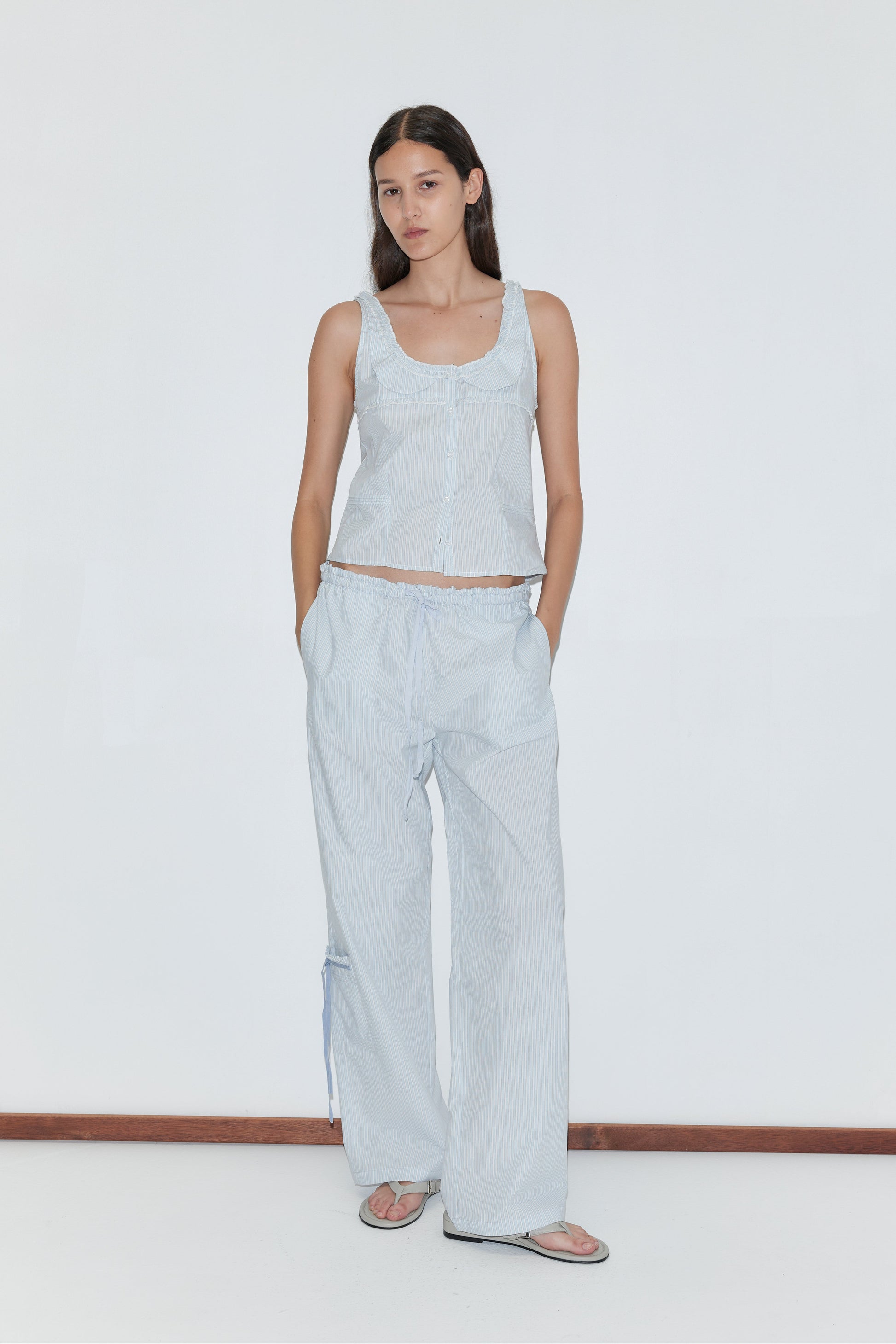 A woman stands against a plain white wall wearing the Pintuck Collar Set - Mist Stripe, featuring a light blue top and matching wide-leg pants. She has her hands in her pockets, pairs the set with sandals, and maintains a neutral expression.