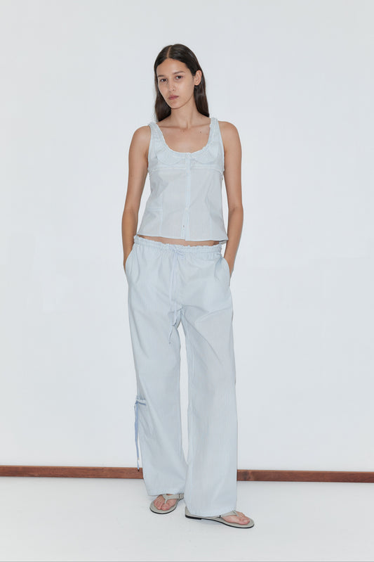 A woman stands against a plain white wall wearing the Pintuck Collar Set - Mist Stripe, featuring a light blue top and matching wide-leg pants. She has her hands in her pockets, pairs the set with sandals, and maintains a neutral expression.