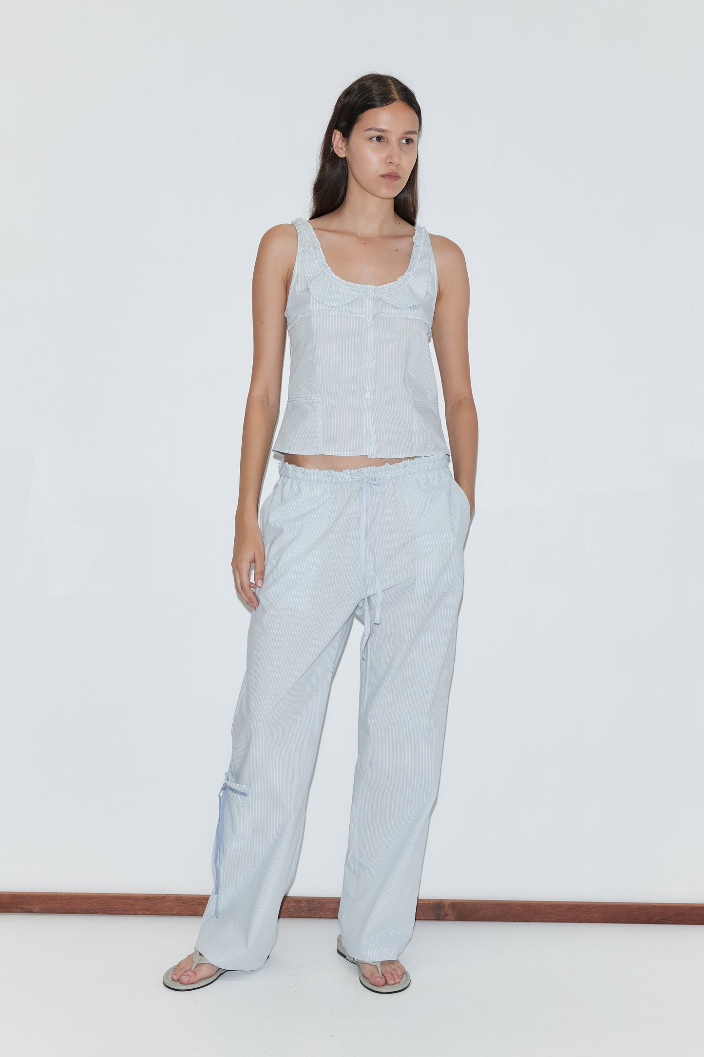 A woman wears the Deiji Studios Frills Top in Mist Stripe with loose drawstring pants, flip-flops, and a neutral expression, standing against a plain white background and looking slightly to the side.