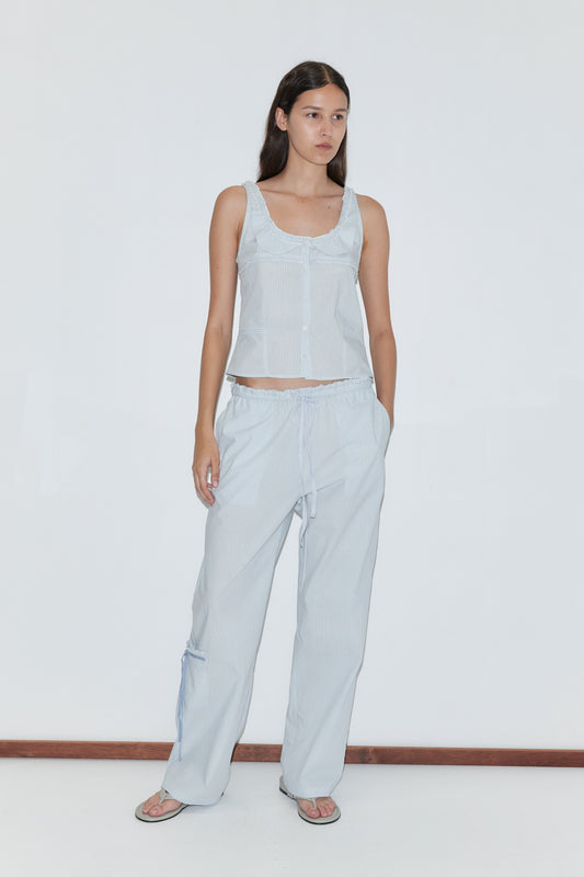 A woman wears the Deiji Studios Frills Top in Mist Stripe with loose drawstring pants, flip-flops, and a neutral expression, standing against a plain white background and looking slightly to the side.