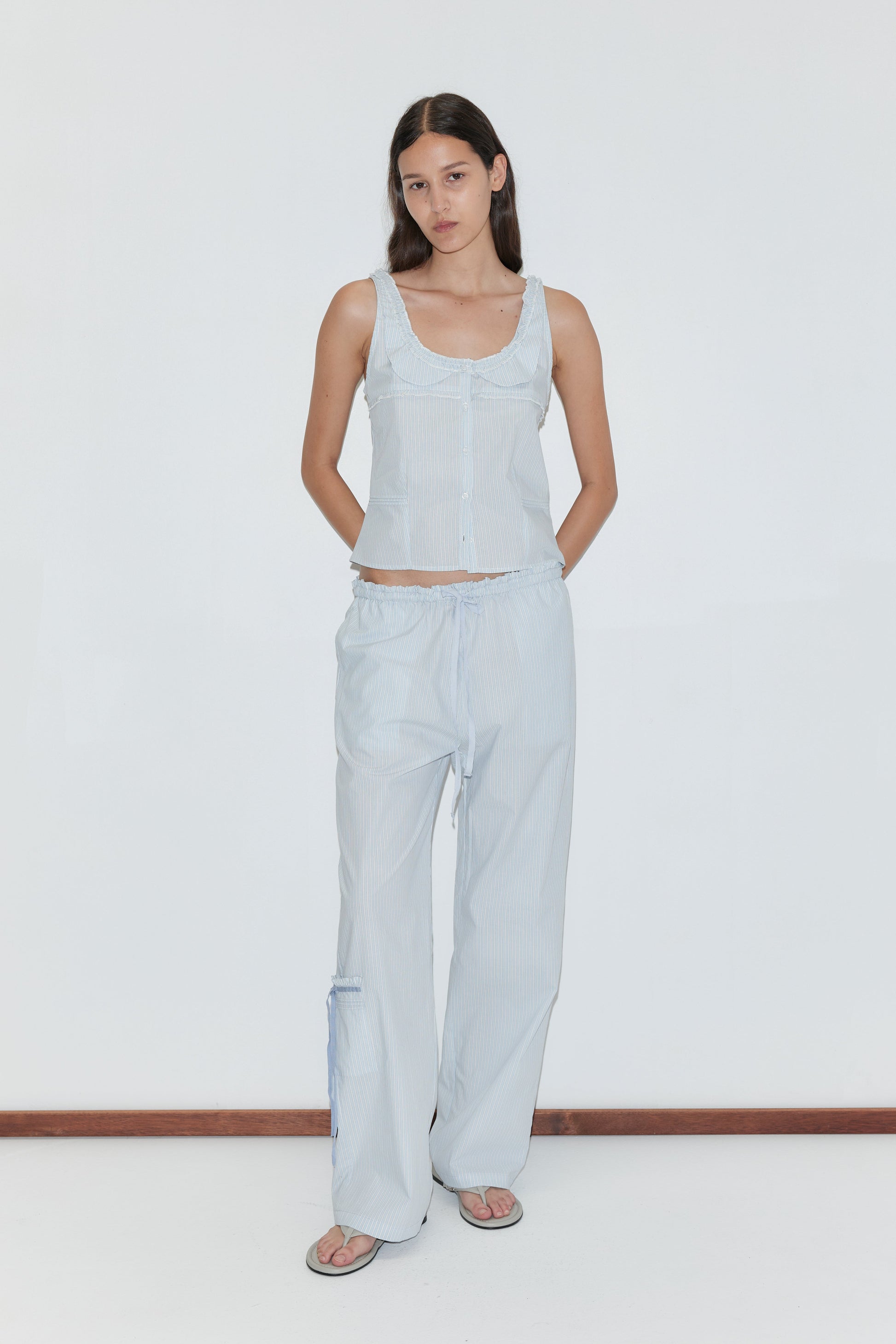 A woman with long dark hair stands against a white background, wearing the Frills Top - Mist Stripe by Deiji Studios with matching loose pants and sandals. Her hands are behind her back.