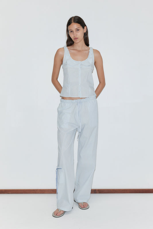 A woman with long dark hair stands against a white background, wearing the Frills Top - Mist Stripe by Deiji Studios with matching loose pants and sandals. Her hands are behind her back.