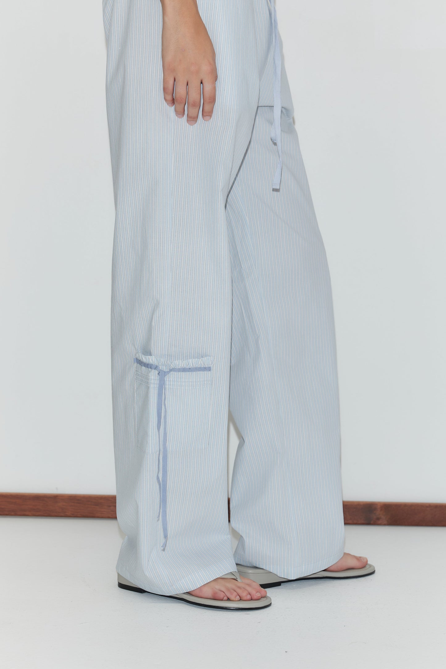A person wearing the Pintuck Collar Set in Mist Stripe—featuring wide-leg, light blue and white striped pants—stands on a white floor against a white wall. Only the lower half of their body is visible.
