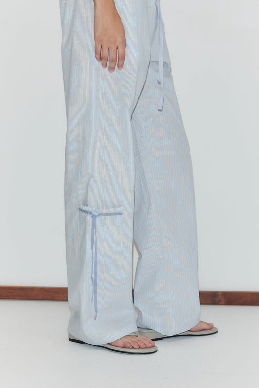 A person wearing the Pintuck Collar Set in Mist Stripe—featuring wide-leg, light blue and white striped pants—stands on a white floor against a white wall. Only the lower half of their body is visible.
