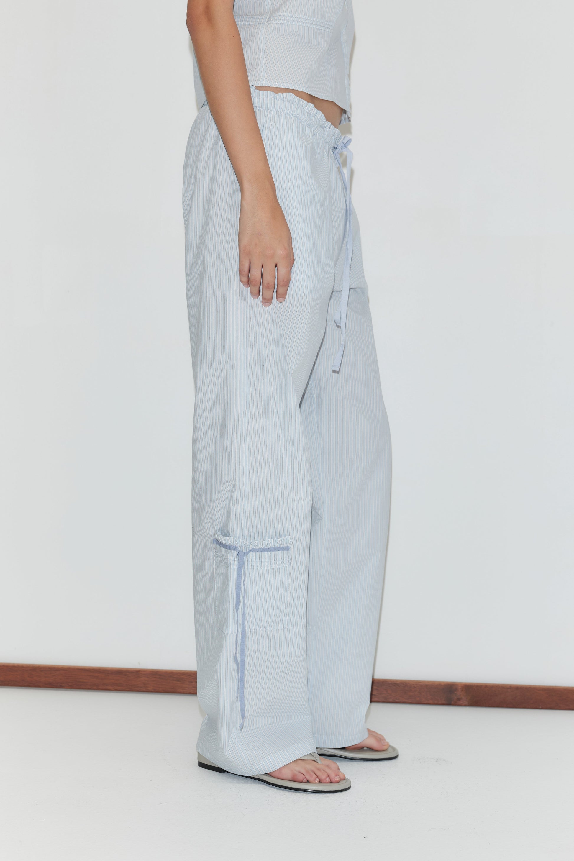 A person stands sideways against a white background wearing the Grosgrain Pant - Mist Stripe, featuring light blue relaxed legs, subtle vertical stripes, and an elastic waistband, paired with a matching top and black flip-flops.