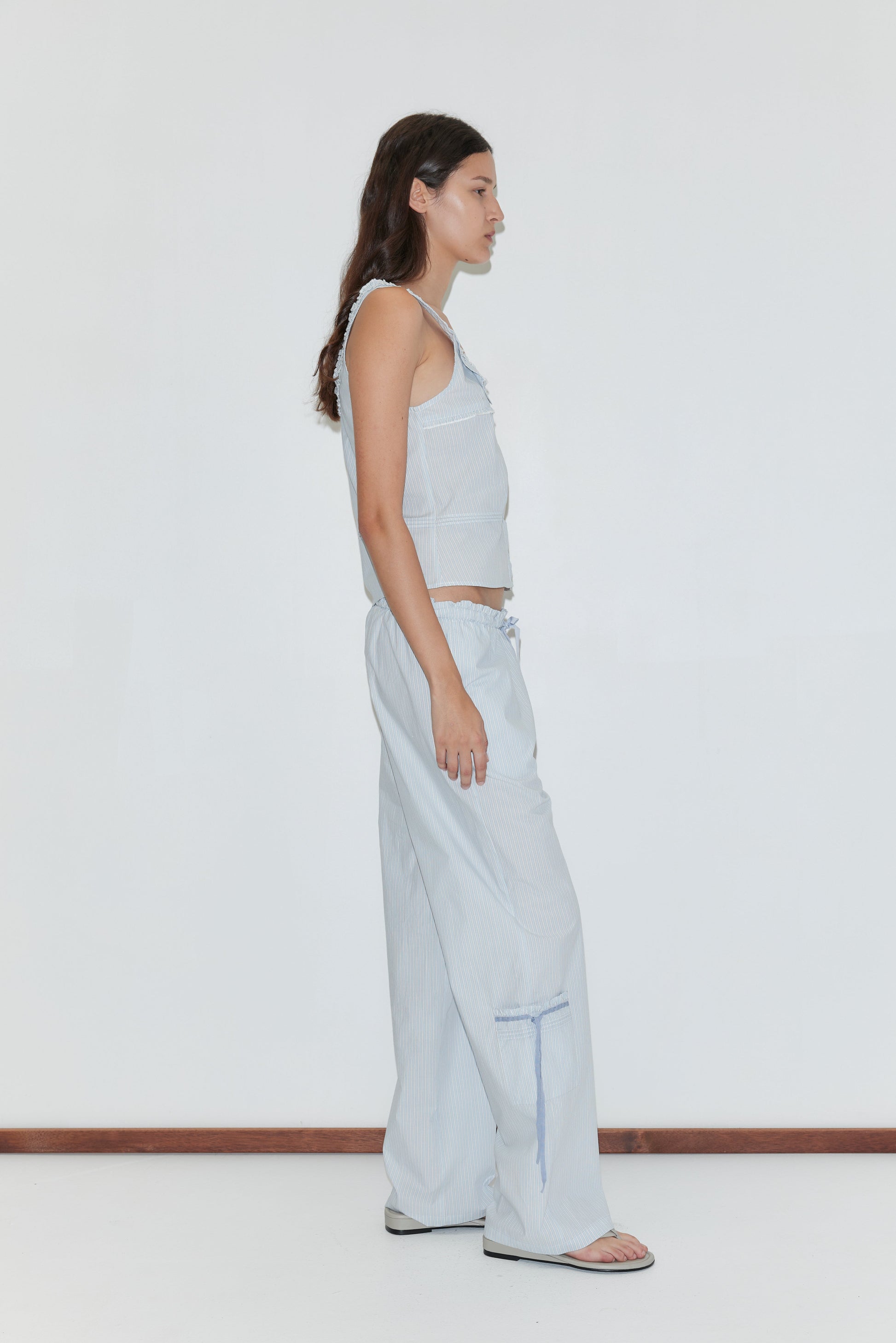 A woman stands in profile against a white background, wearing the Frills Top - Mist Stripe by Deiji Studios with matching wide-leg drawstring pants and sandals.