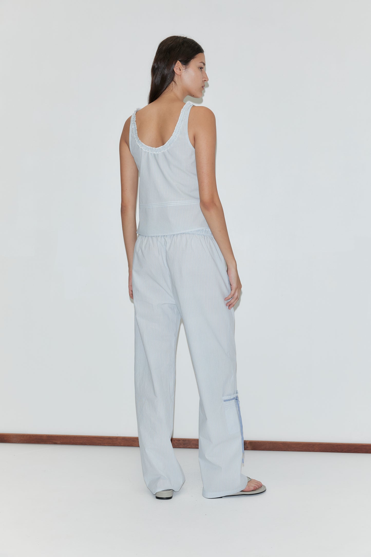 A woman stands barefoot on a white floor against a plain background, wearing the Grosgrain Pant - Mist Stripe: light blue relaxed leg pants with an elasticated waistband and subtle geometric stripes. She faces away with long dark hair.