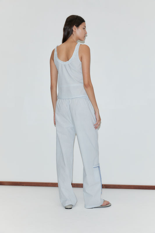 A woman stands barefoot on a white floor against a plain background, wearing the Grosgrain Pant - Mist Stripe: light blue relaxed leg pants with an elasticated waistband and subtle geometric stripes. She faces away with long dark hair.