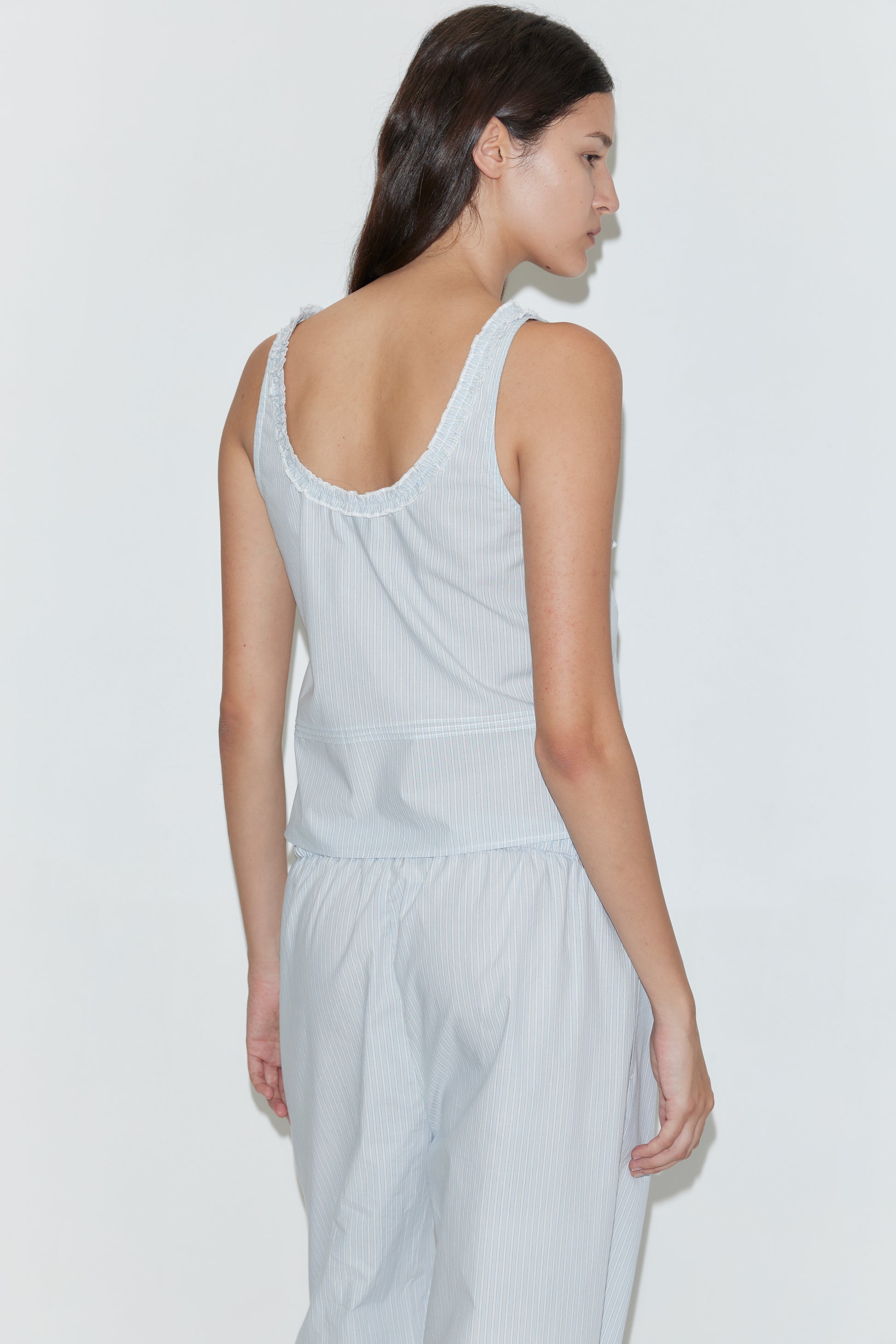 A woman with long brown hair stands sideways, wearing the Frills Top - Mist Stripe by Deiji Studios and matching light blue and white striped pants. The relaxed-fit, sleeveless cotton poplin outfit is set against a plain white background.