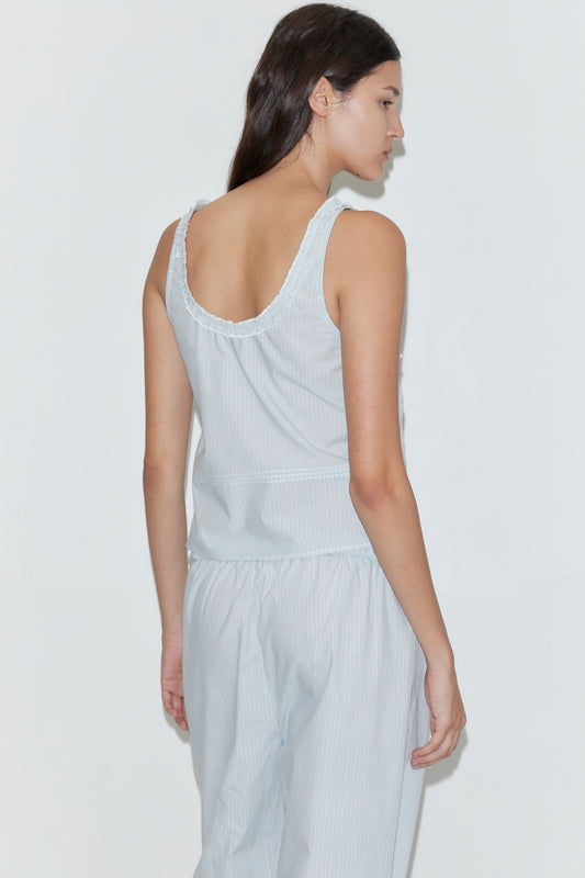 A woman with long brown hair stands sideways, wearing the Frills Top - Mist Stripe by Deiji Studios and matching light blue and white striped pants. The relaxed-fit, sleeveless cotton poplin outfit is set against a plain white background.