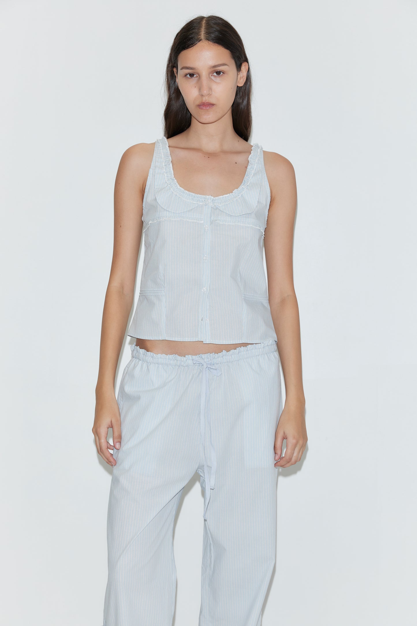 A woman with long dark hair wears the Grosgrain Pant in Mist Stripe, featuring a relaxed fit and elasticated waistband, paired with a light blue sleeveless top. She stands against a plain white background, looking directly at the camera.