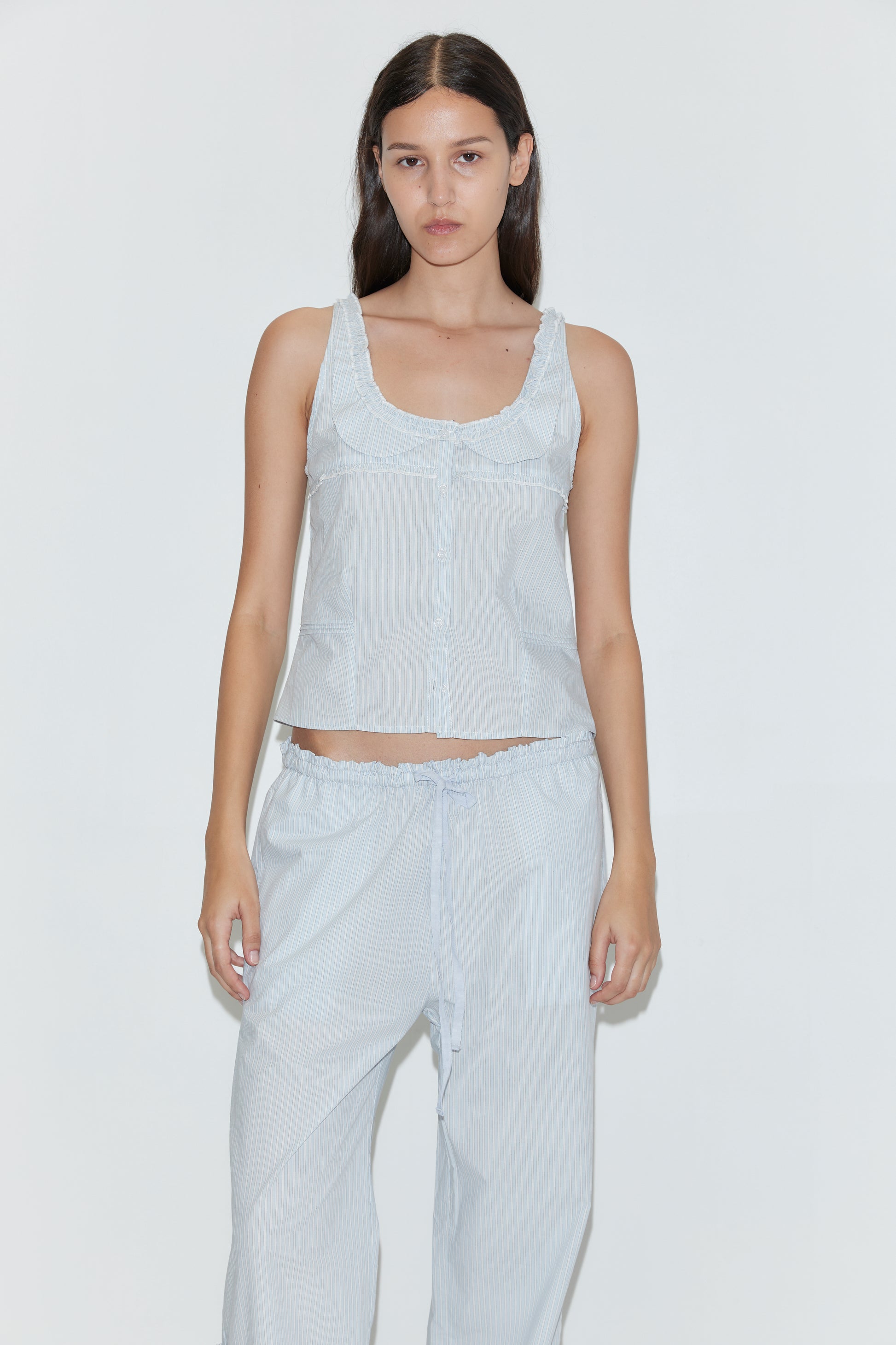A woman with long dark hair wears the Grosgrain Pant in Mist Stripe, featuring a relaxed fit and elasticated waistband, paired with a light blue sleeveless top. She stands against a plain white background, looking directly at the camera.