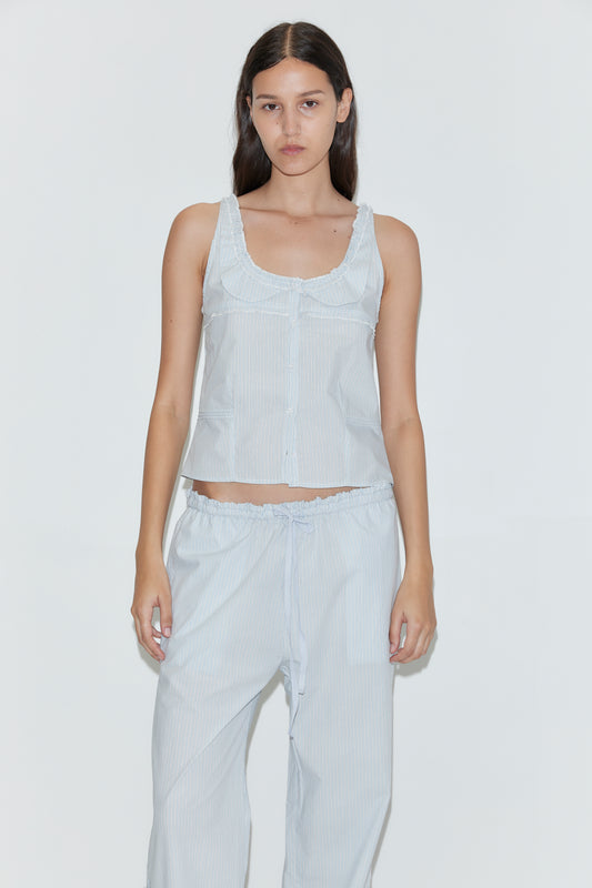 A woman with long dark hair wears the Grosgrain Pant in Mist Stripe, featuring a relaxed fit and elasticated waistband, paired with a light blue sleeveless top. She stands against a plain white background, looking directly at the camera.