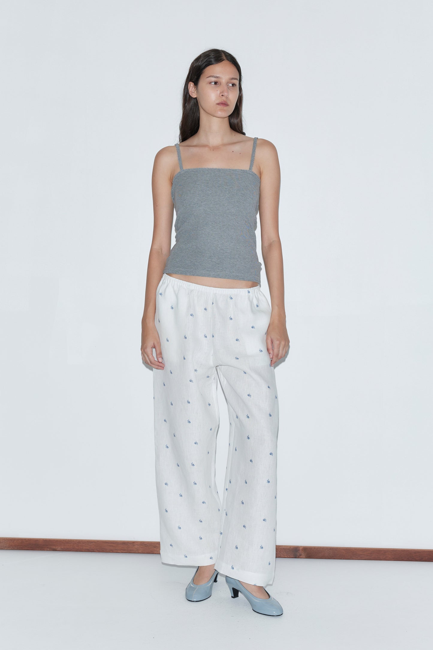 A woman with long dark hair and a neutral expression stands against a plain white background wearing the Linen Ease Trouser - Floral Blue, a gray spaghetti strap top, and light blue heels.