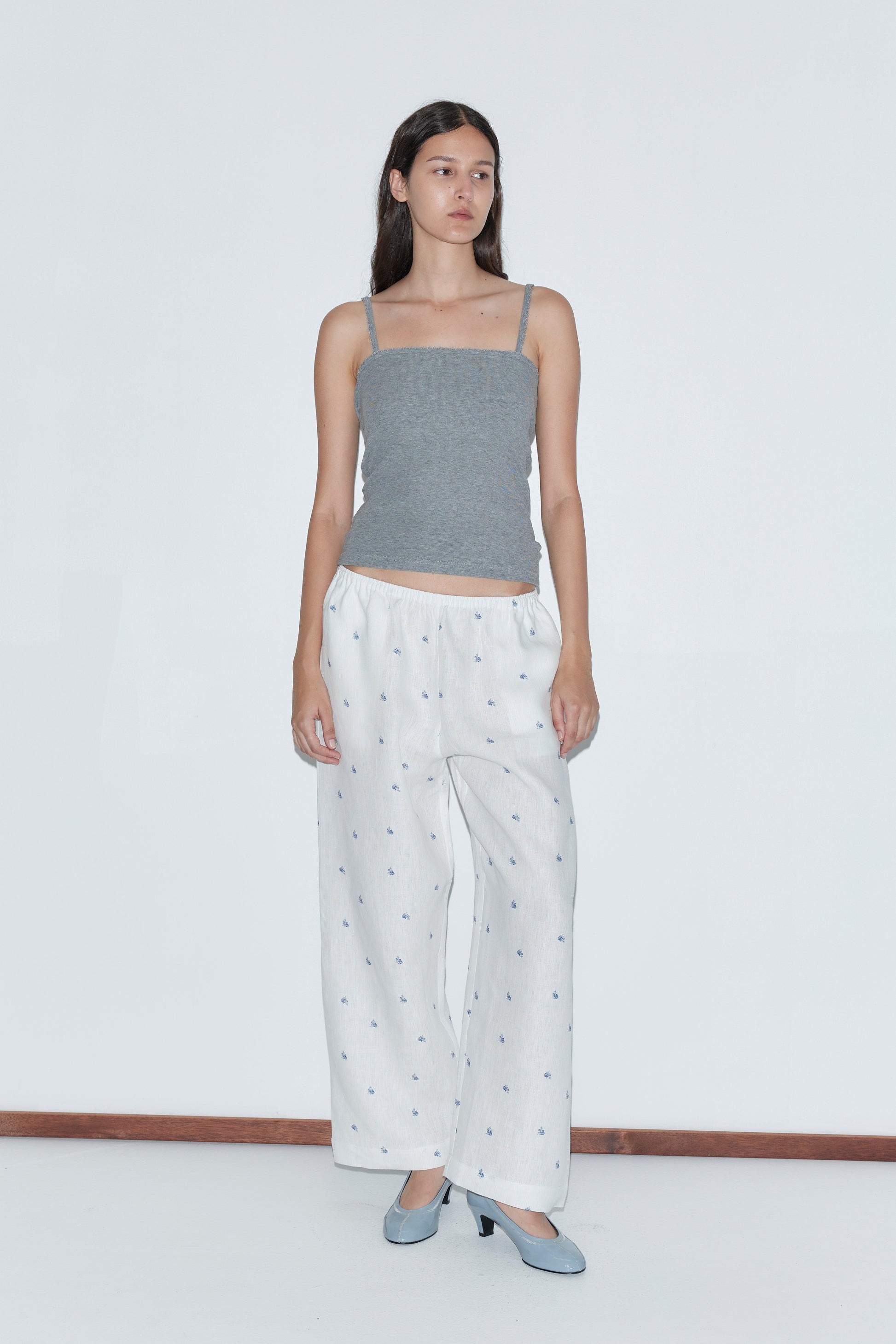 A woman with long dark hair and a neutral expression stands against a plain white background wearing the Linen Ease Trouser - Floral Blue, a gray spaghetti strap top, and light blue heels.
