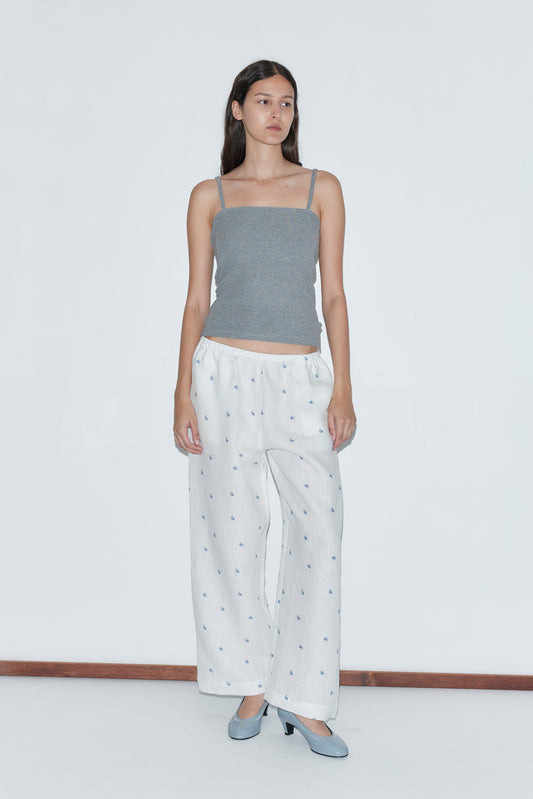 A woman with long dark hair and a neutral expression stands against a plain white background wearing the Linen Ease Trouser - Floral Blue, a gray spaghetti strap top, and light blue heels.