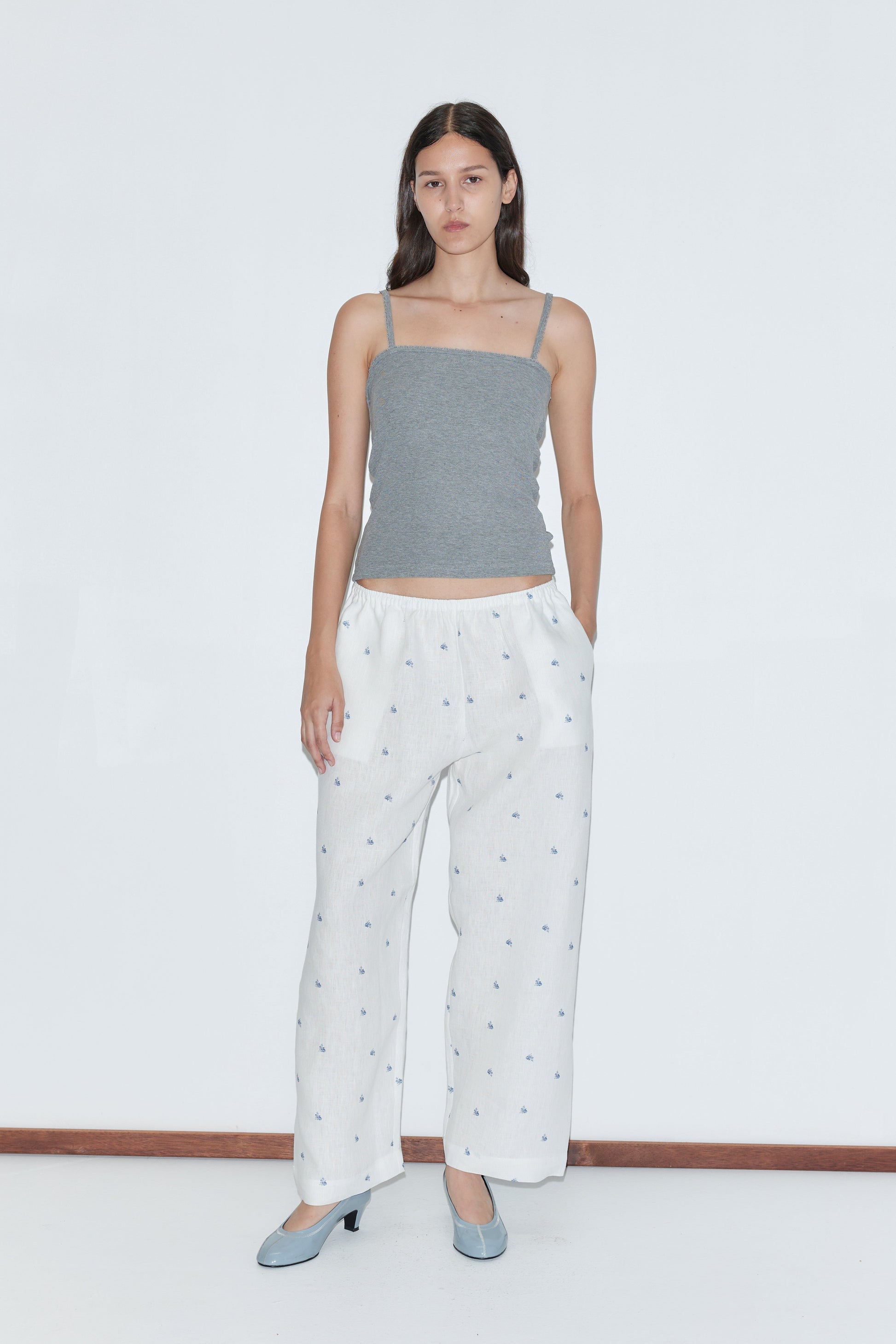 A woman stands against a white background, wearing gray spaghetti straps and the Linen Ease Trouser - Floral Blue. She pairs the loose, elastic-waist floral blue trousers with light blue heels, hands in her pockets and a neutral expression.