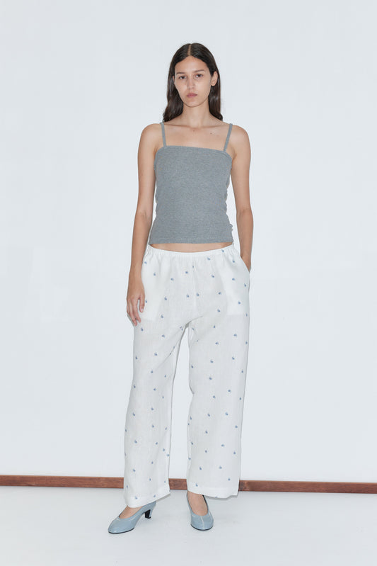 A woman stands against a white background, wearing gray spaghetti straps and the Linen Ease Trouser - Floral Blue. She pairs the loose, elastic-waist floral blue trousers with light blue heels, hands in her pockets and a neutral expression.