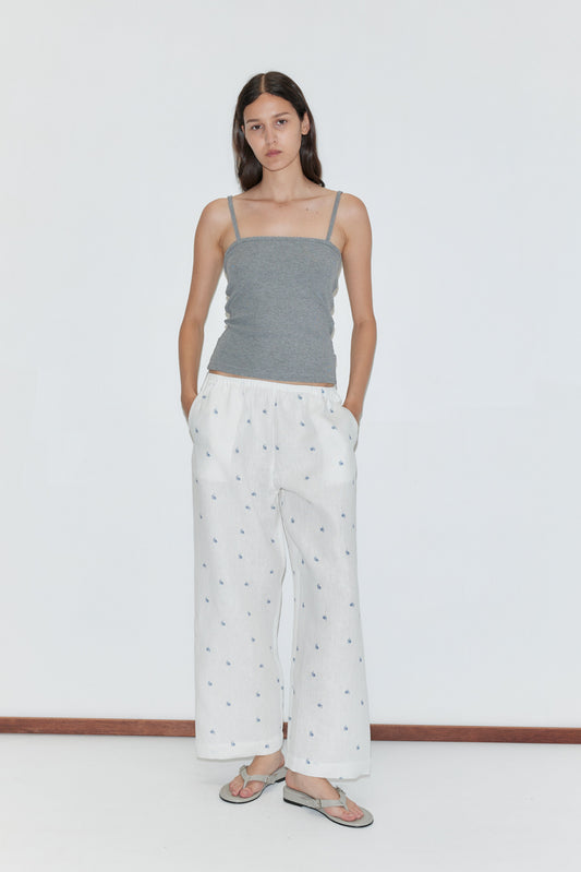 A woman with straight brown hair stands against a plain white background, wearing the Linen Ease Trouser—loose white pants with a subtle print—paired with a gray spaghetti strap top and sandals, her hands in her pockets.