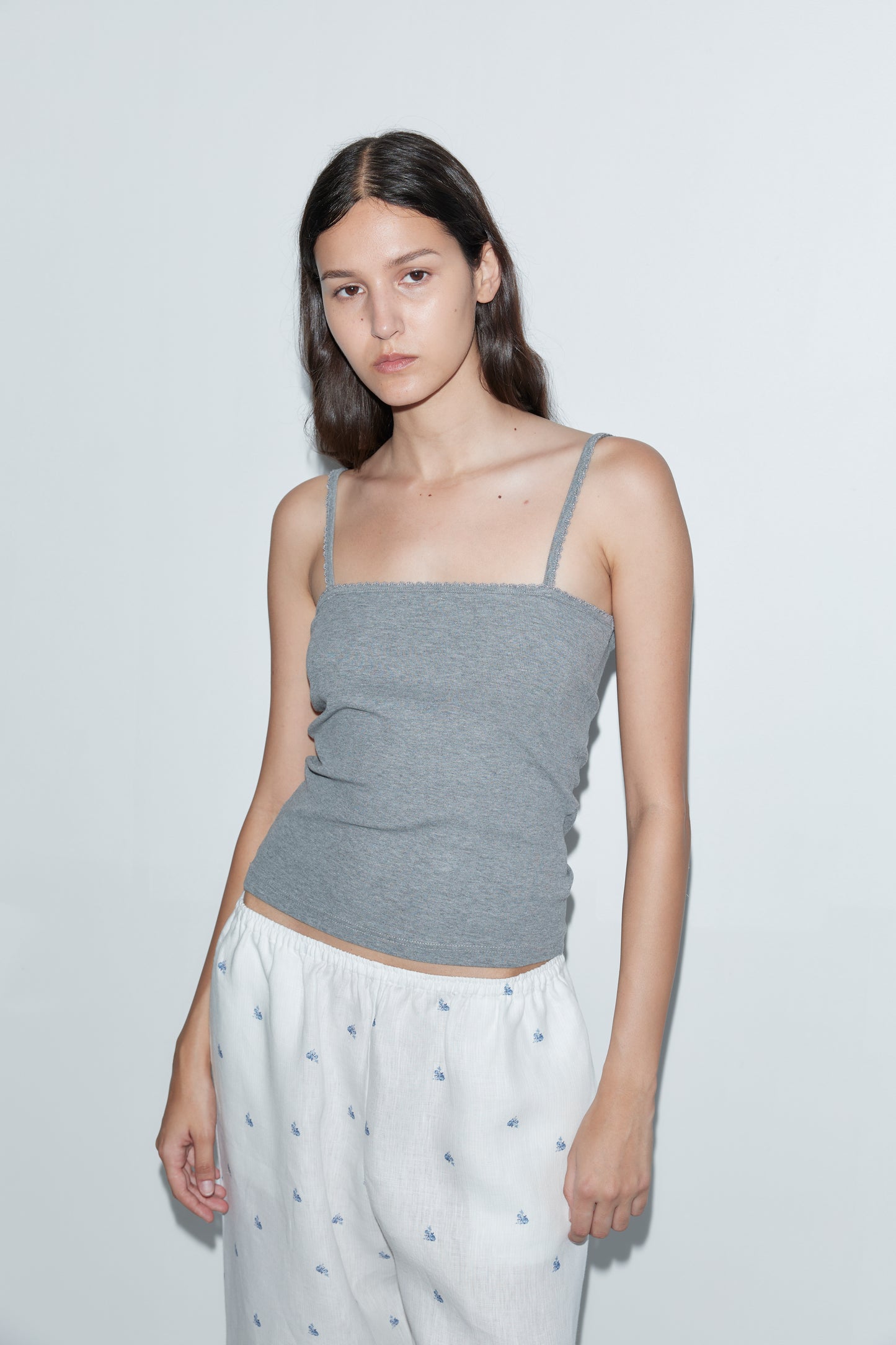 A young woman with long dark hair stands against a plain white wall, wearing a gray spaghetti strap top and the Linen Ease Trouser - Floral Blue. She has a neutral expression and her arms rest relaxed at her sides.
