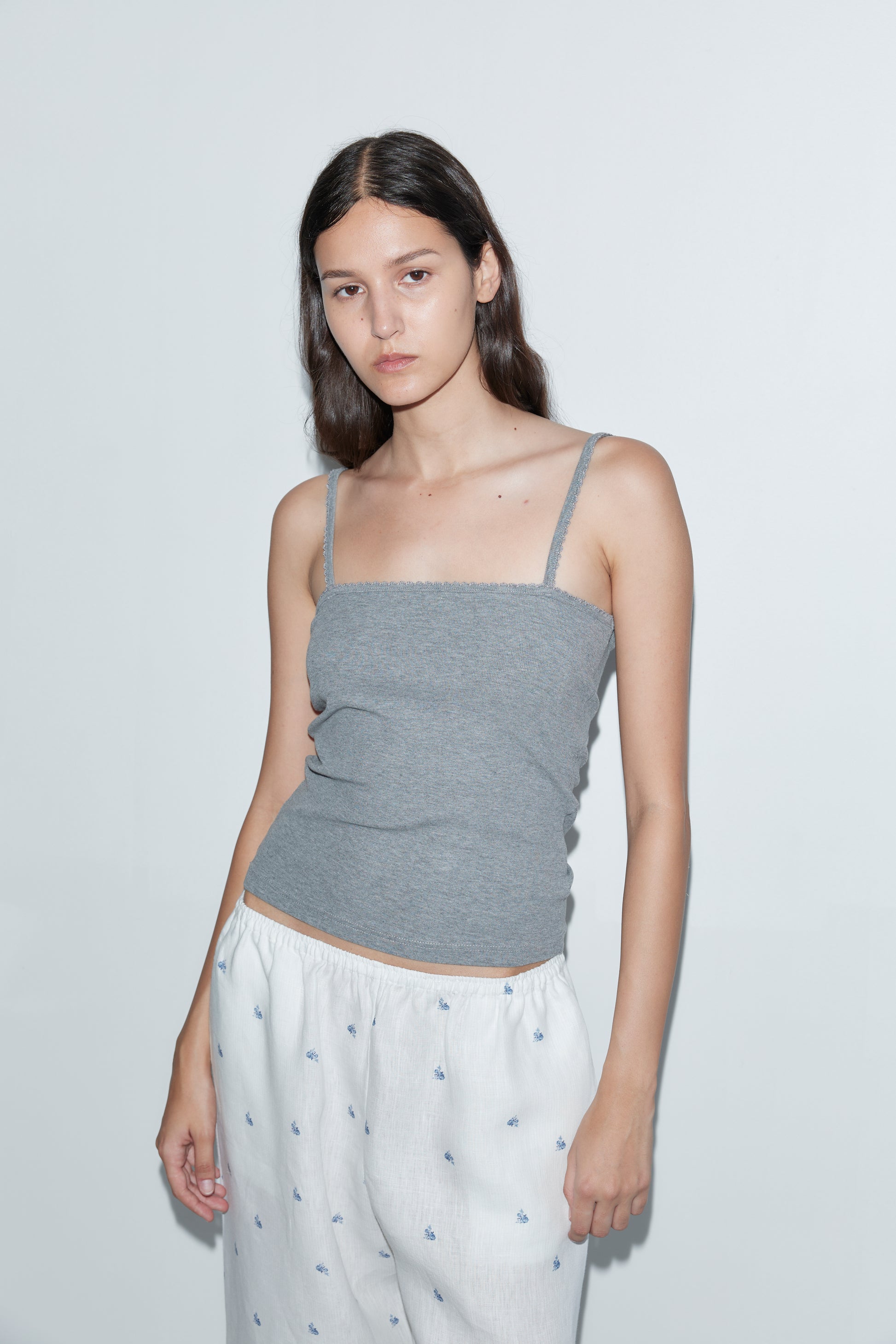 A young woman with long dark hair stands against a plain white wall, wearing a gray spaghetti strap top and the Linen Ease Trouser - Floral Blue. She has a neutral expression and her arms rest relaxed at her sides.