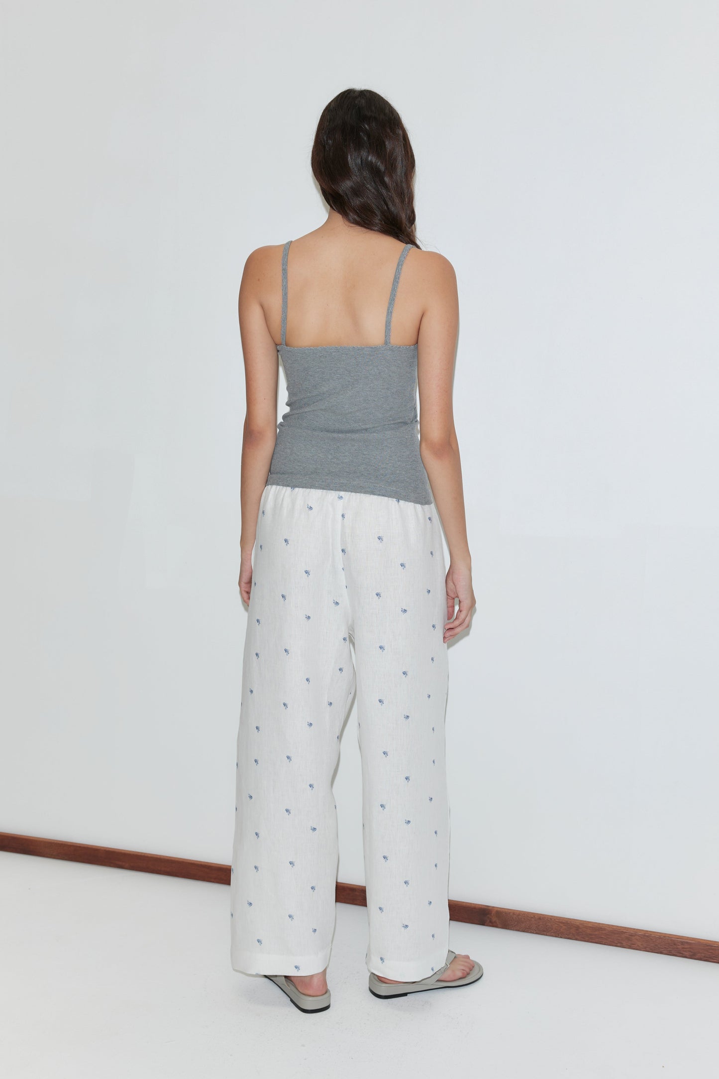 A woman stands facing away from the camera, wearing a gray camisole, sandals, and the Linen Ease Trouser - Floral Blue with an elastic waist and floral blue pattern, against a plain white background.