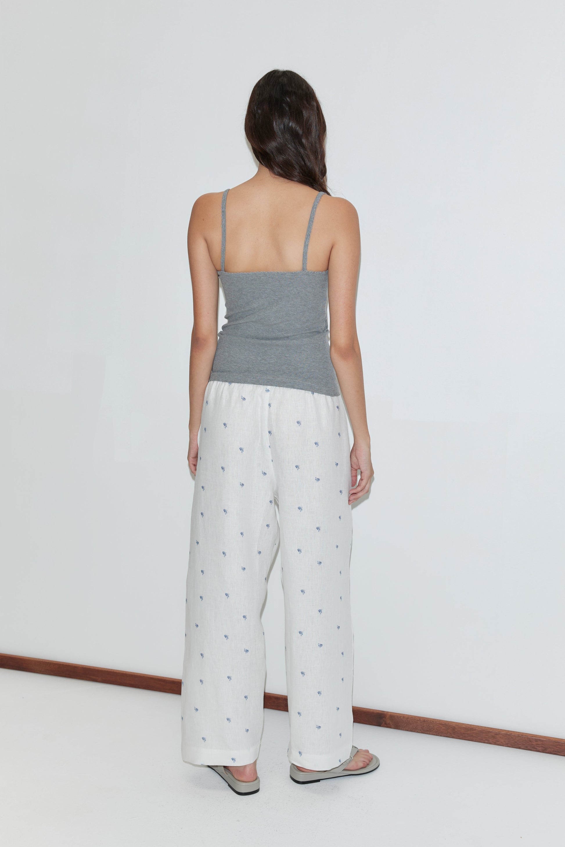 A woman stands facing away from the camera, wearing a gray camisole, sandals, and the Linen Ease Trouser - Floral Blue with an elastic waist and floral blue pattern, against a plain white background.
