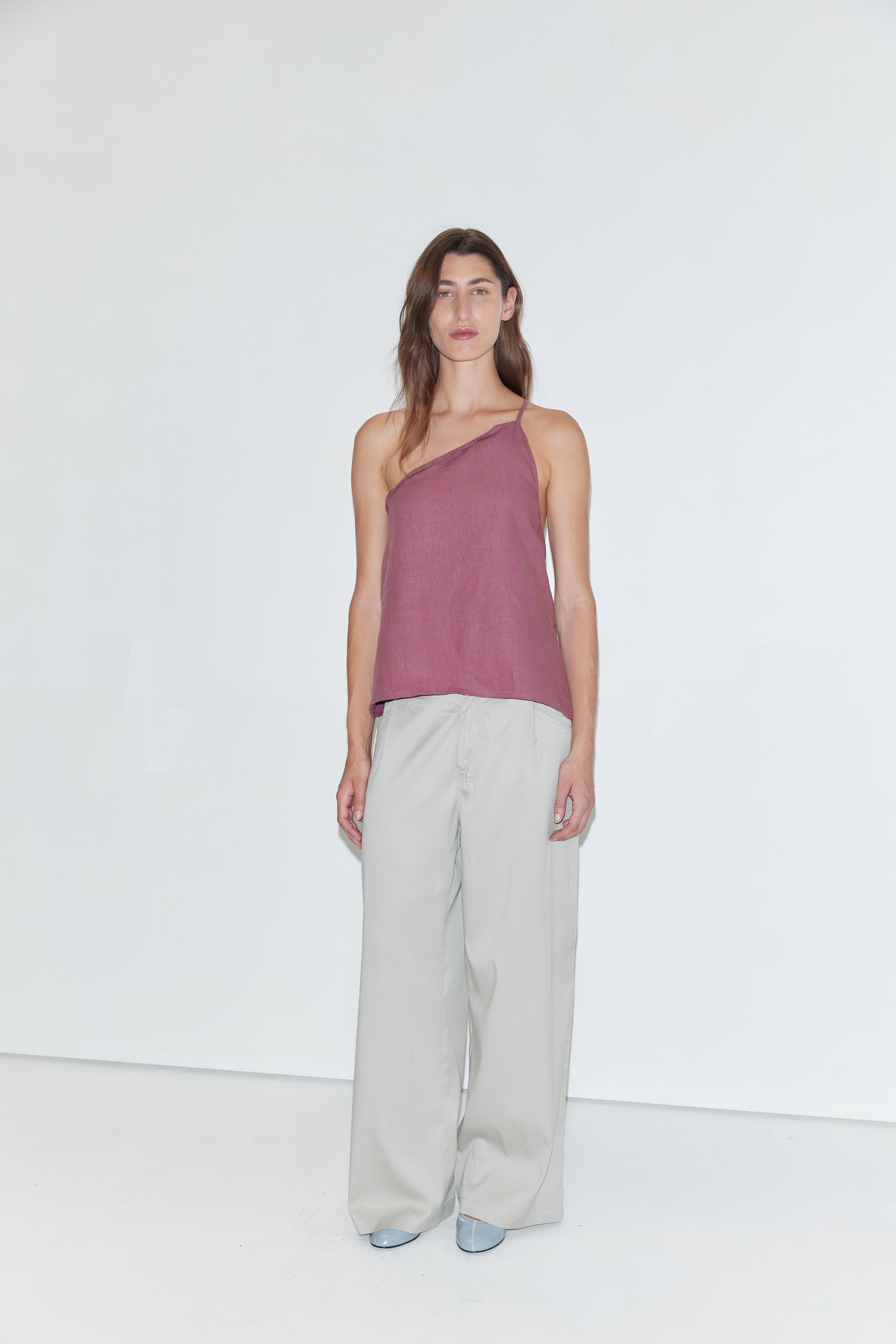 A person stands against a plain white background wearing the Half Halter - Brick linen one-shoulder top from Deiji Studios, paired with loose light gray pants and light blue shoes.