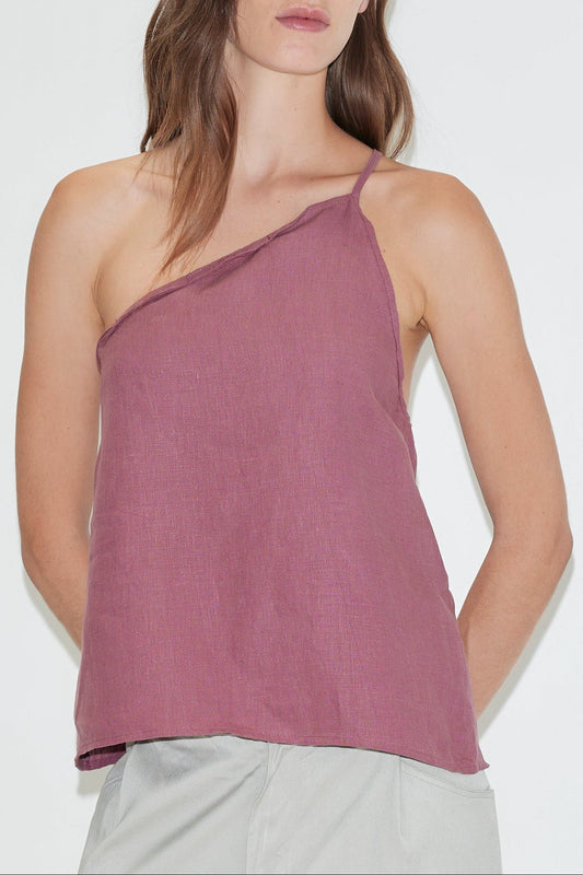A woman wearing the Half Halter - Brick, a sleeveless linen one-shoulder top from Deiji Studios, paired with light-colored pants and standing against a plain white background.