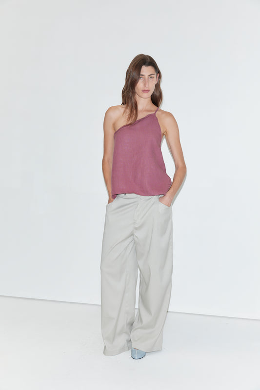 A woman stands against a white background, wearing the Half Halter - Brick linen one-shoulder top from Deiji Studios, wide-legged light gray pants, and blue shoes, with her hands in her pockets.